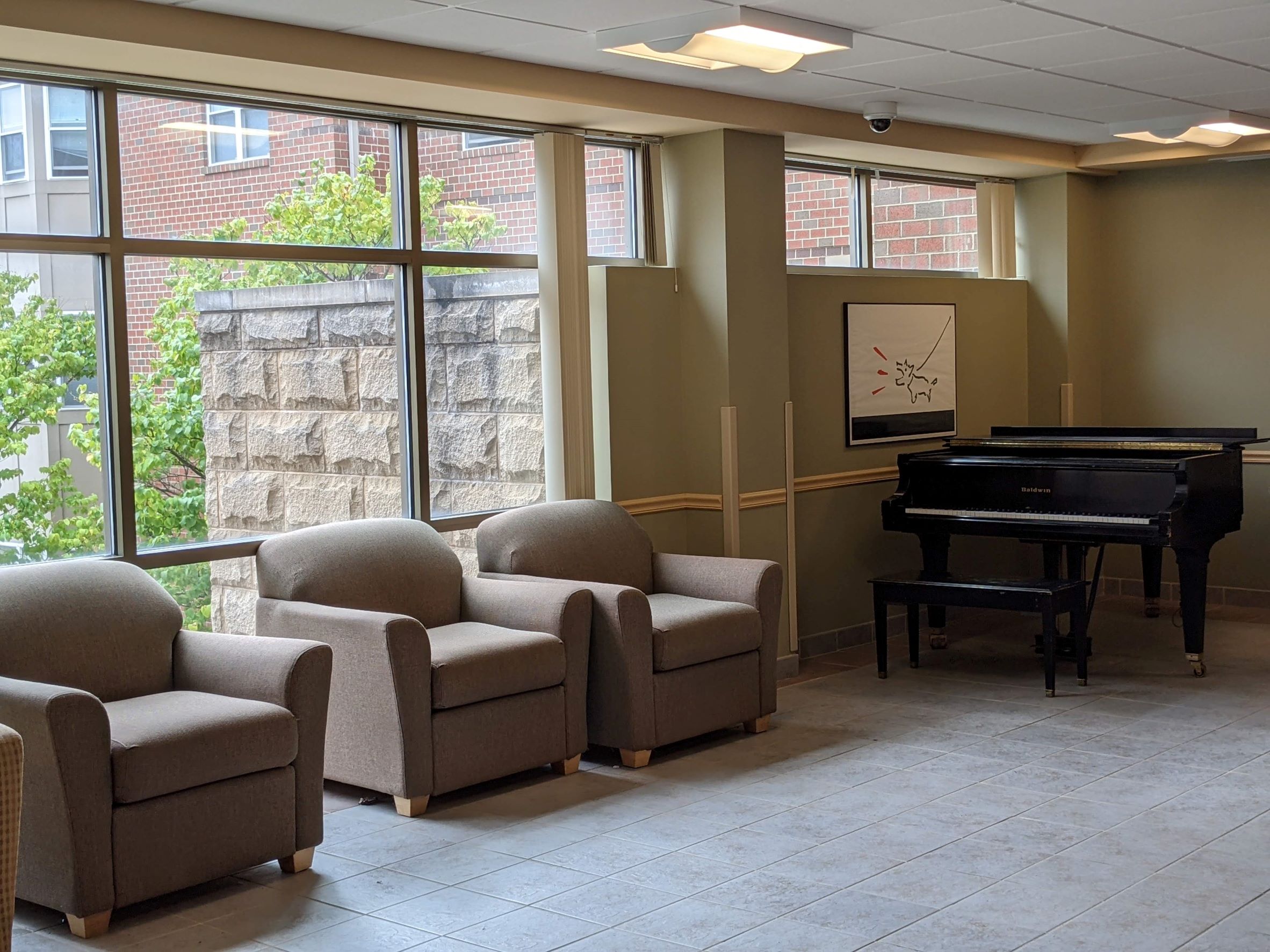 Living-Learning Dormitory Community lounge with large cushy chairs and a baby grand piano in the background with a large glass window and stone wall with vegetation beyond