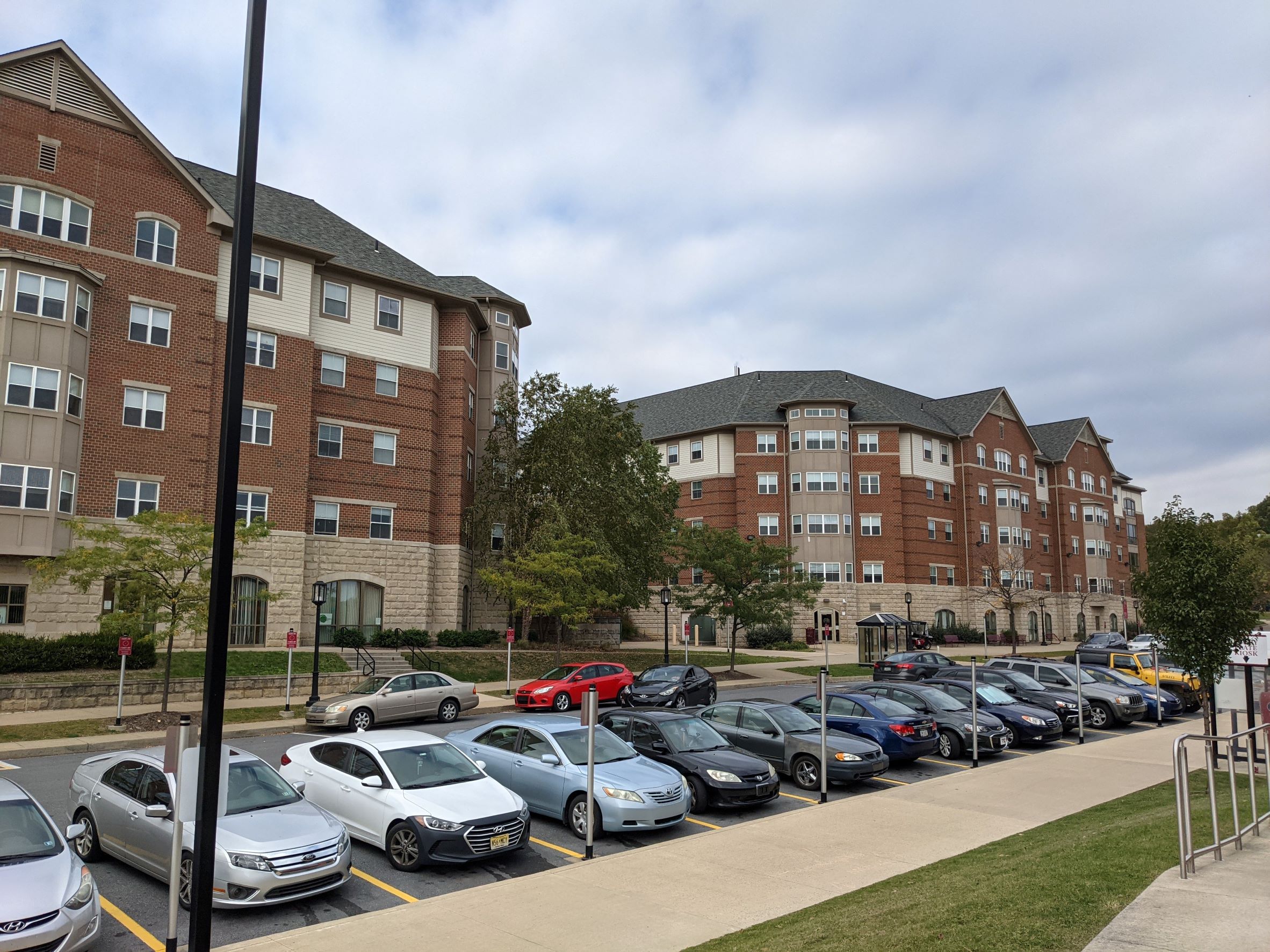 Traditional looking five story red brick  living learning student housing community dormitory building with clapboard siding and large gables and a large stone base with storefront windows in it along a busy road with many cars parked