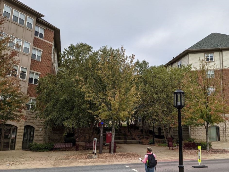 Large sidewalk and ramping system zig-zagging up a small hillside on campus in a densely wooded area between two living learning student housing community dormitory buildings, woman crossing street in foreground with streetlight