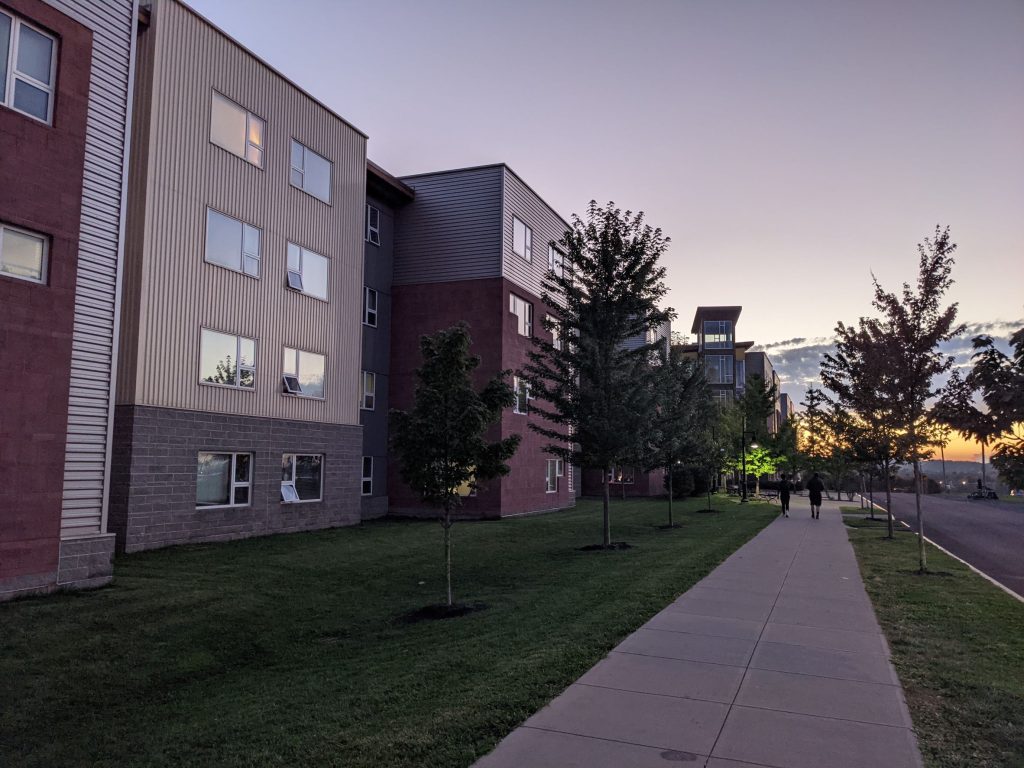 series of disconnected smaller student housing buildings along a walkway with a large tower in the distance set against the backdrop of a beautiful sunset